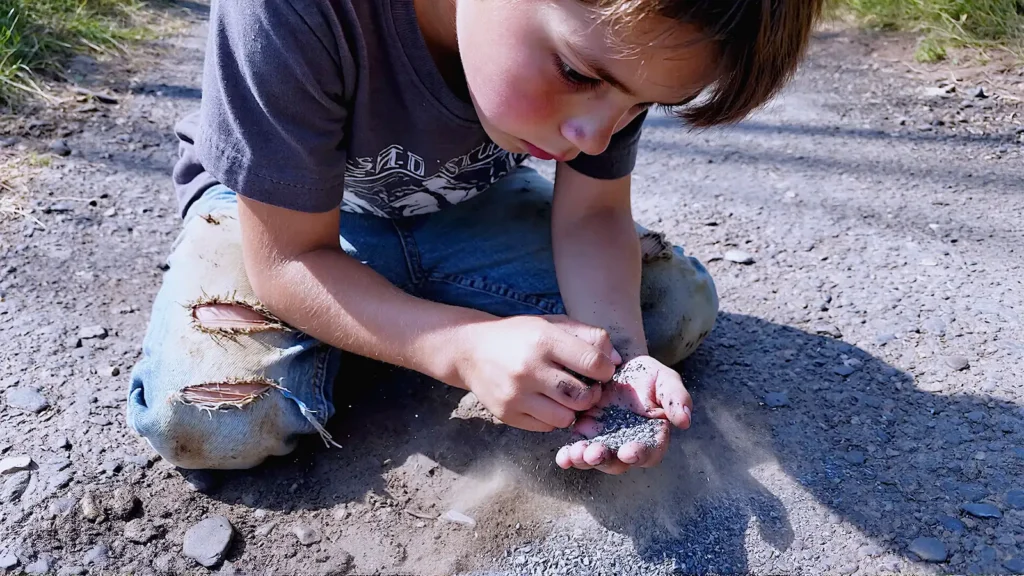 A cinematic, photorealistic close-up low angle shot of a 7-year-old child getting up from a fall on a rough asphalt path. The child is on one knee, brushing dirt off their palms. The expression is not tearful, but focused and assessing. Natural sunlight, harsh shadows, realistic textures (scuffed denim, gravel, dust). 35mm film grain style, Kodak Portra 400. No adults in the frame.