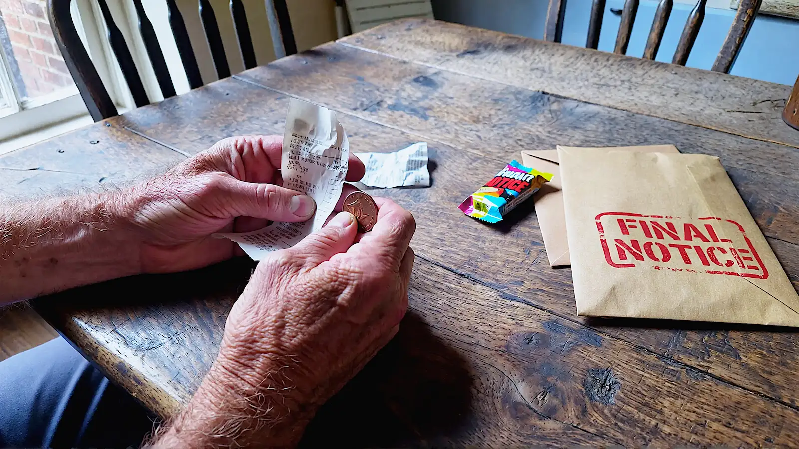 A close-up, eye-level shot of a weathered British kitchen table made of dark oak. Action: A pair of rough, working hands (middle-aged, calloused) are holding a very small, crinkled grocery receipt and a single copper penny. Context: On the table, a stark contrast: a modern, brightly coloured but noticeably small chocolate bar wrapper next to a generic brown envelope with a "FINAL NOTICE" red stamp. Lighting: Natural, moody window light coming from the left, casting long shadows. It’s an overcast UK afternoon (cool blue tones in the shadows). Style: Cinematic realism, Kodak Portra 400 film stock style. High texture—you can feel the wood grain and the paper quality. No cartoons, no floating 3D icons. Mood: Somber, grounded, resilient, but tired.